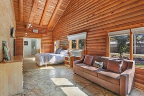 A log cabin interior with a stone floor, wooden walls, a bed, a sofa, and a window with a view of the outdoors. - WLCRS - Unit 4 -Walk to Market Days -  Resort Pool! (Wimberley)