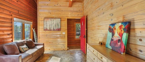 A log cabin interior with a stone floor, wooden walls, a brown leather sofa, a wooden dresser, and a painting of a cow.