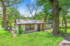 A small, single-story house with a stone foundation and a metal roof, surrounded by lush greenery and trees. - The Guadalupe River right in your backyard! River living at its best! (Wimberley)