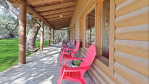 A covered porch with wooden beams, a row of red chairs, and a view of a grassy area with trees. - WLCRS - Unit 4 -Walk to Market Days -  Resort Pool! (Wimberley)