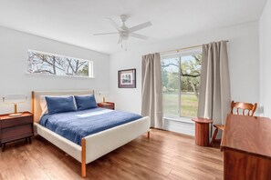 Un dormitorio con cama, ventilador de techo, ventana con cortinas, mesita de noche, una silla y una mesa de madera.
