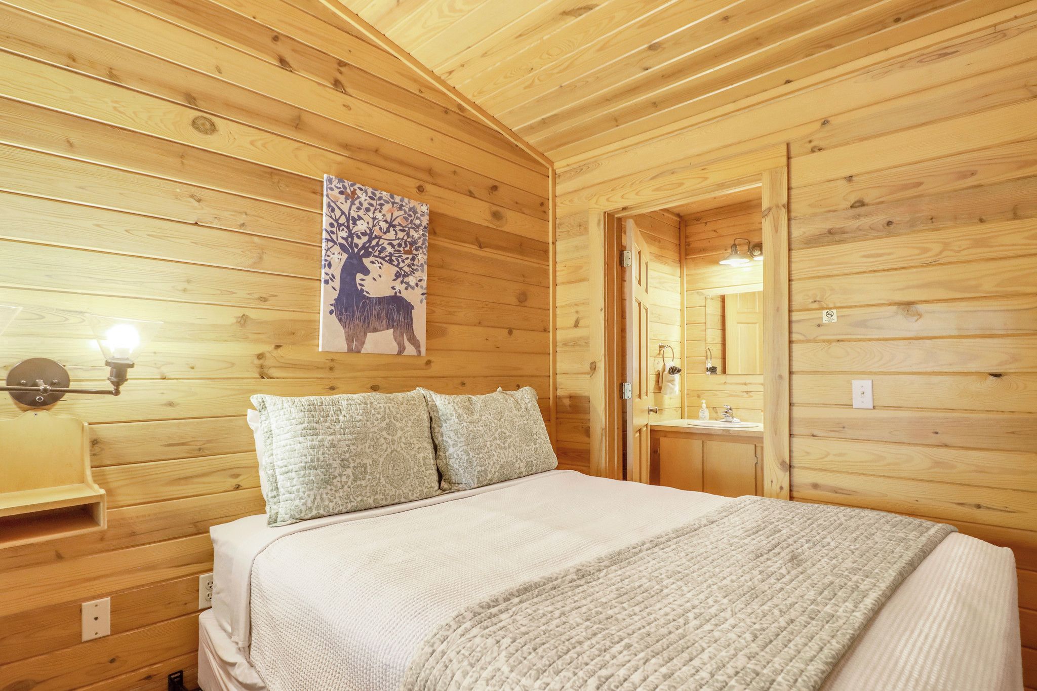 A wooden cabin bedroom with a bed, two pillows, a nightstand, a wall-mounted lamp, and a bathroom visible through an open door.