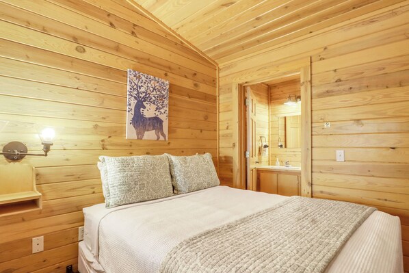 A wooden cabin bedroom with a bed, two pillows, a nightstand, a wall-mounted lamp, and a bathroom visible through an open door.