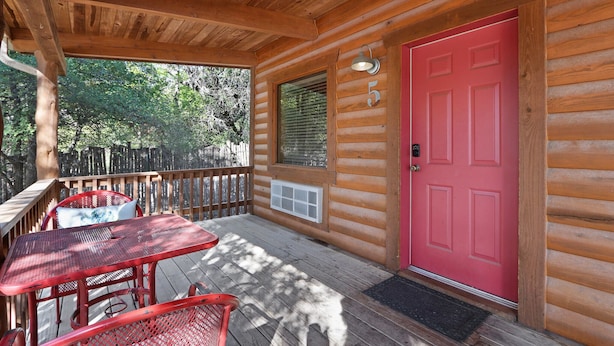 Eine Holzhütte mit roter Tür, einer Veranda mit Tisch und Stühlen sowie einem Fenster mit Jalousien.