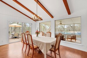Un comedor con una gran mesa, sillas de madera y una araña. Se ven grandes ventanales con vistas a una piscina y árboles.