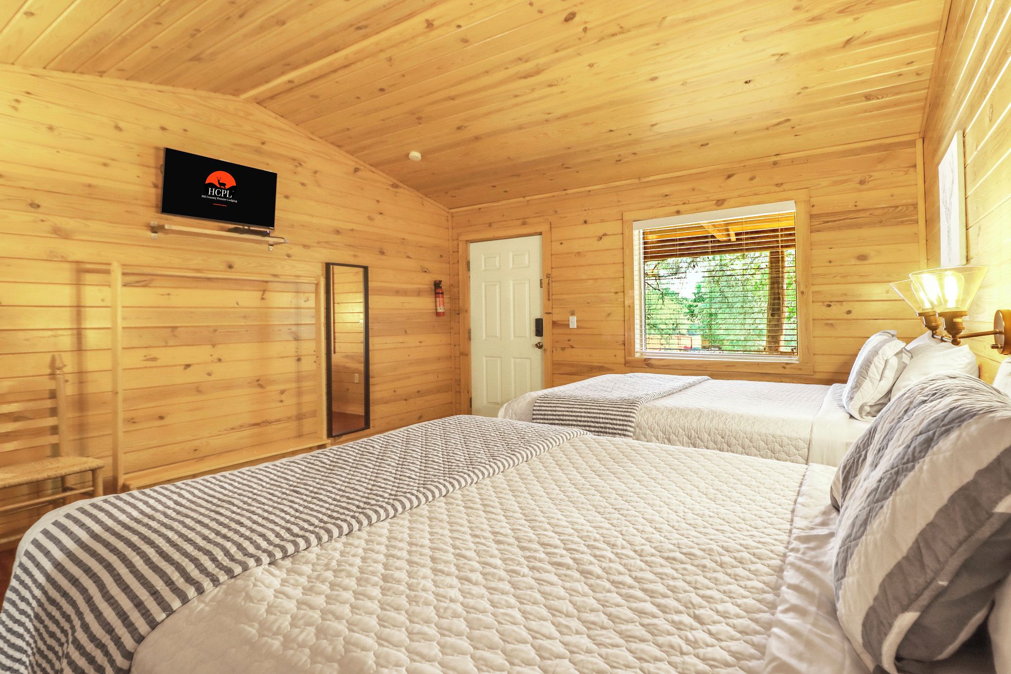 Habitación de cabaña de madera con dos camas, un televisor, una puerta y una ventana con persianas.