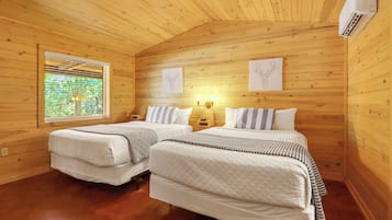 Habitación de cabaña de madera con dos camas, una ventana con persianas, luces fijadas a la pared y obras de arte enmarcadas.