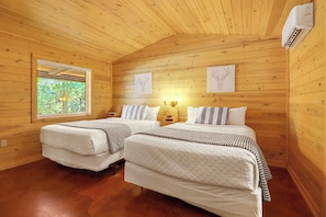 Een houten kamer met twee bedden, een raam met zonwering, wandlampen en kunstwerken in lijsten.