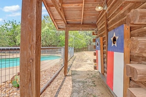 A wooden cabin with a covered porch, a pool, and a fenced area with trees in the background. - Resort Style Swimming Pool, Fire Pit, Grill, and Bocce Ball! (Wimberley)