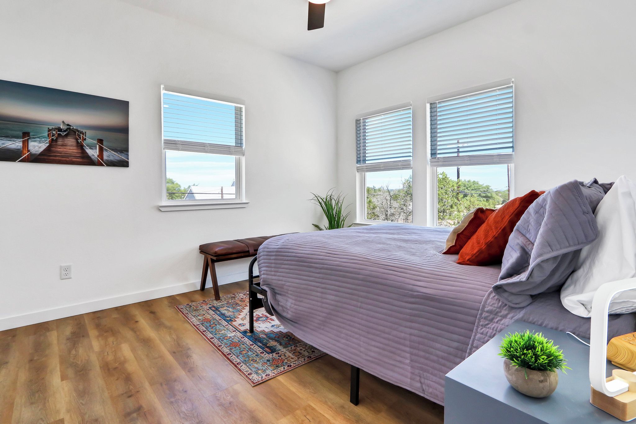 A bedroom with a bed, a bench, a rug, a plant, and a painting on the wall.