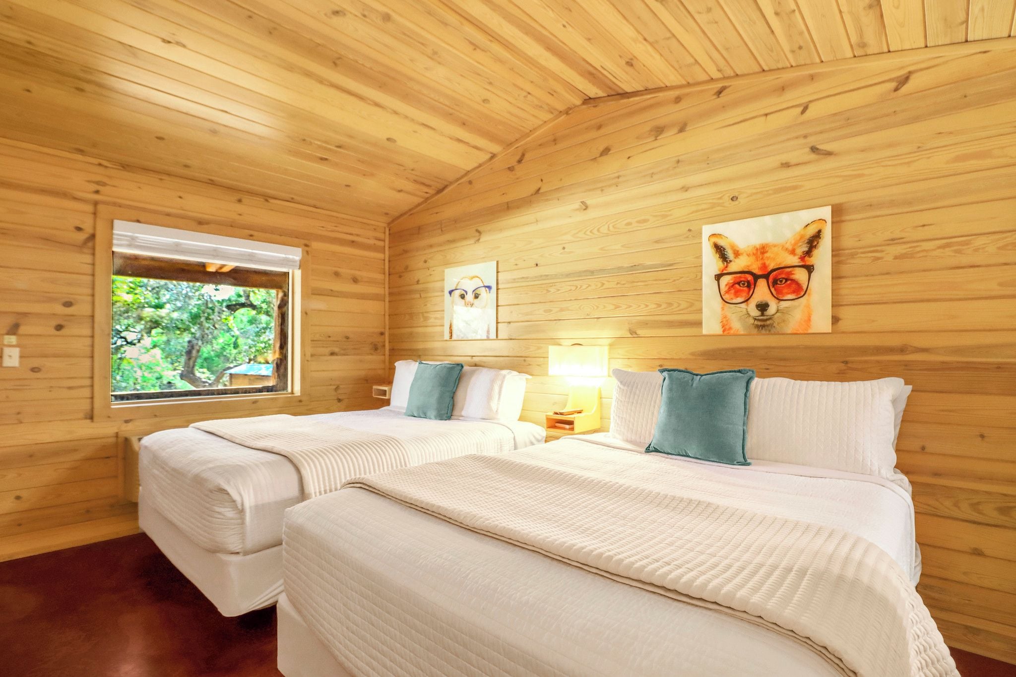 Ein Zimmer in einer Holzhütte mit zwei Betten, einem Fenster mit Blick ins Grüne und zwei an der Wand hängenden Bildern.