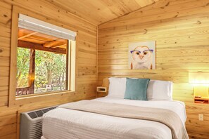 A wooden cabin bedroom with a bed, a window, a painting, and a heater.