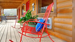 Una cabaña de madera con un porche, una mecedora roja con cojín azul, y una planta grande en maceta.