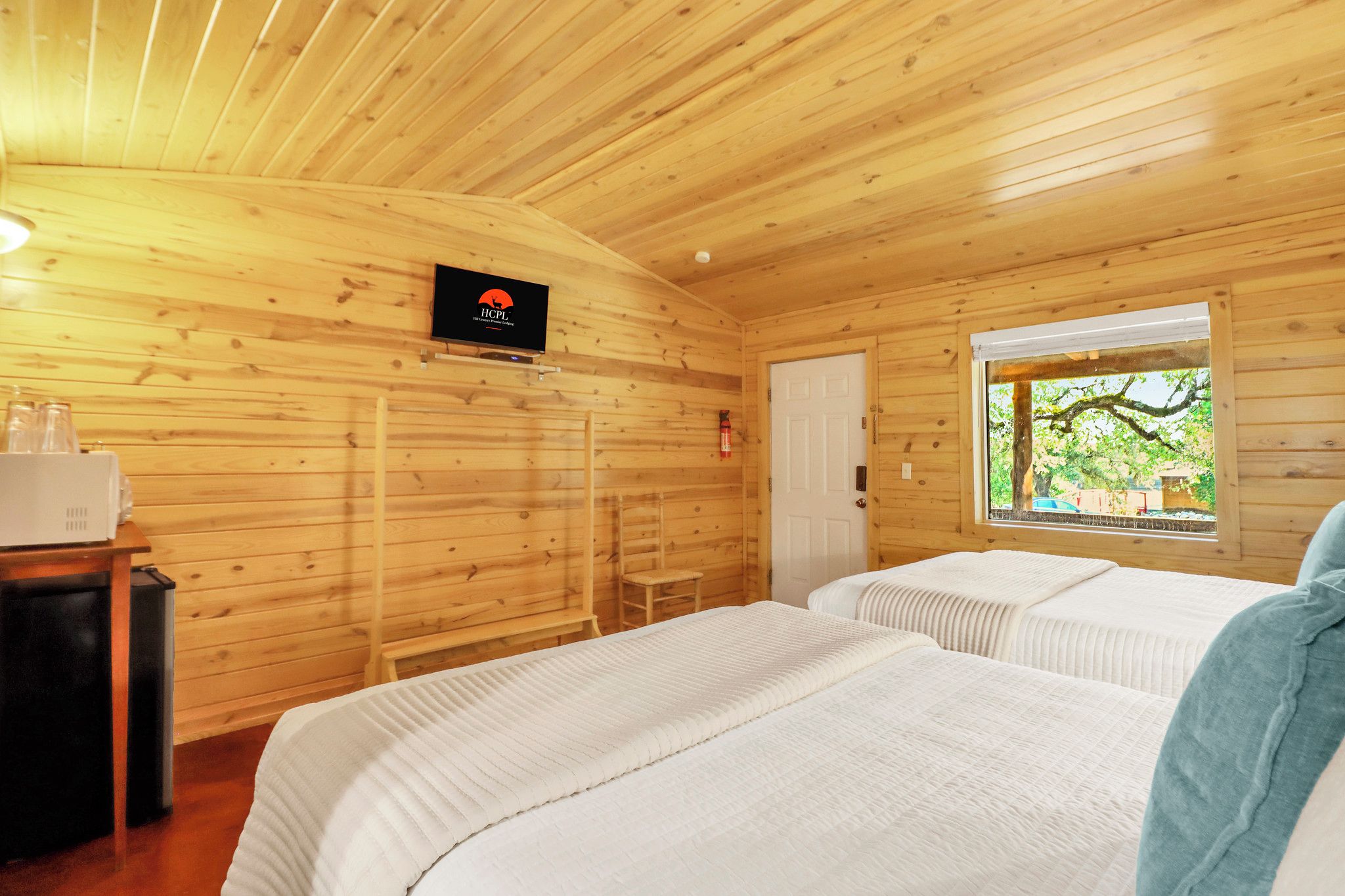 Habitación de cabaña de madera con dos camas, un televisor, un extintor y una ventana con vista a árboles.