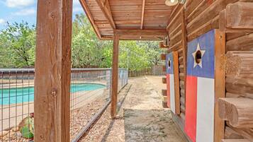 Una cabaña de madera con porche cubierto, piscina y un área cercada con árboles al fondo.
