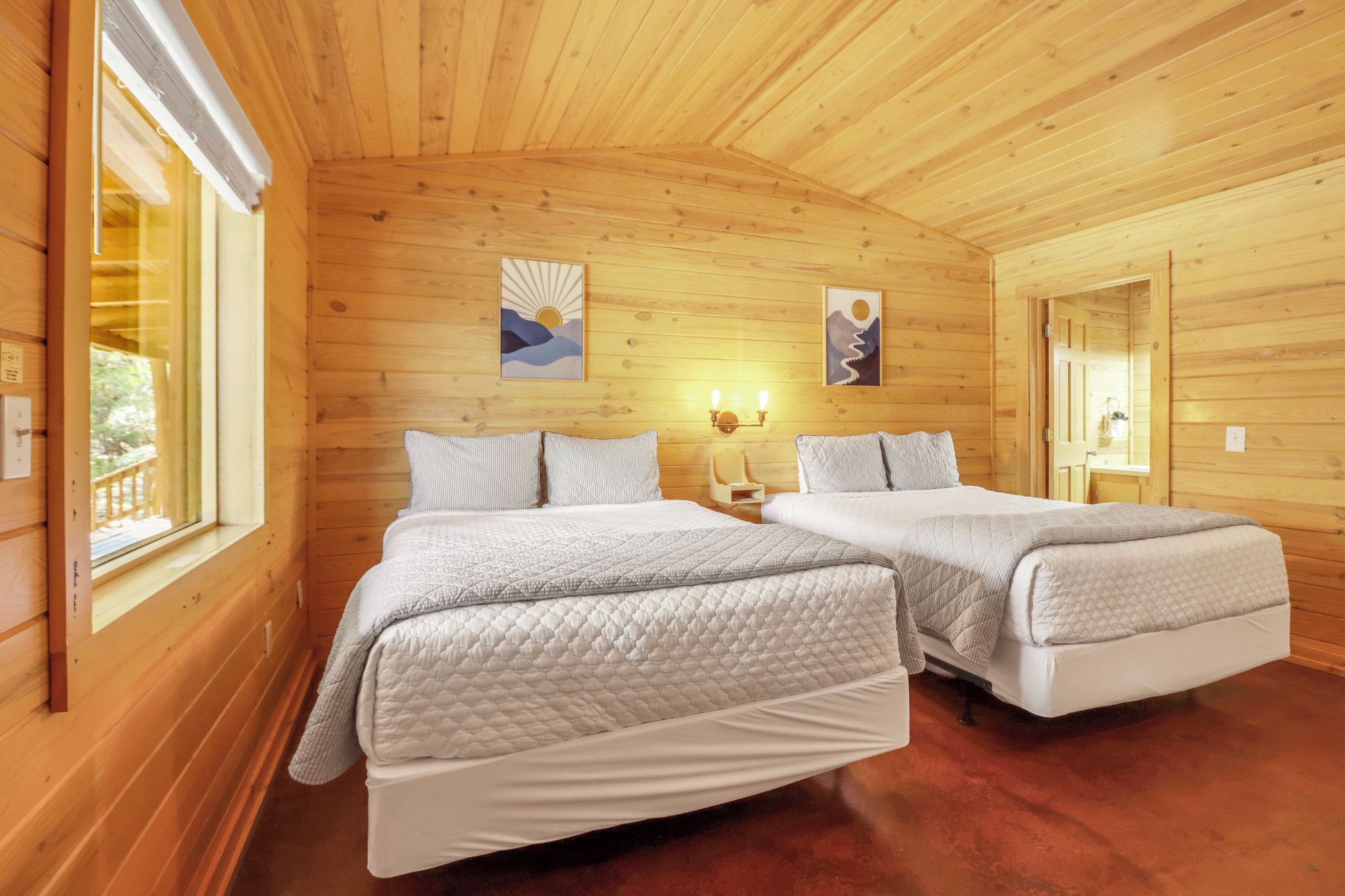 A wooden cabin bedroom with two beds, a window, and artwork on the walls.