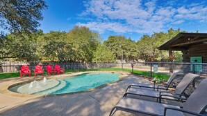 A swimming pool with a fountain, surrounded by red chairs and lounge chairs, with a wooden cabin in the background.