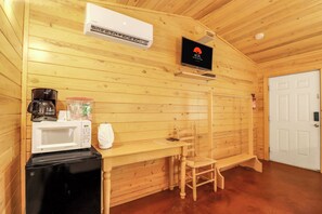 Habitación compacta con detalles de madera, una puerta blanca, una pequeña mesa con una silla, microondas, cafetera y televisor montado en la pared.