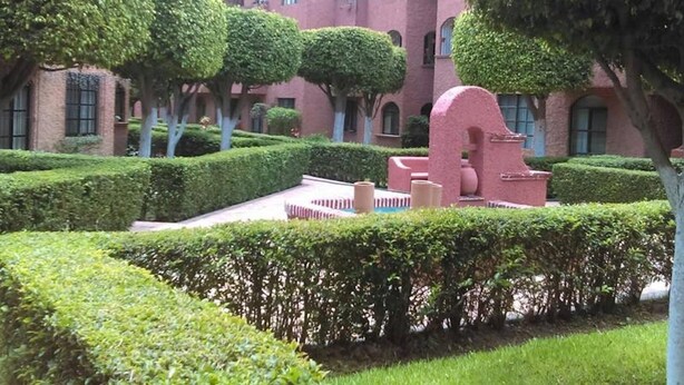 A courtyard with neatly trimmed hedges, a central sculptural element, and a building with arched windows and a terracotta roof. - Departamento Comodo, Centrico, Estacionamiento (Querétaro)