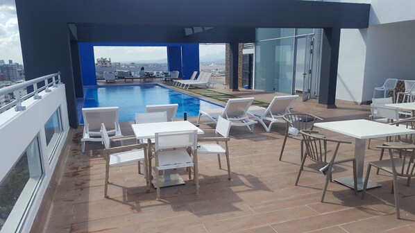 Zona de piscina en la azotea con sillas y mesas de descanso blancas, vistas despejadas de la ciudad y un diseño arquitectónico moderno.