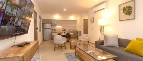 A modern living room with a sofa, coffee table, and TV. There is a kitchen in the background with cabinets and a refrigerator.