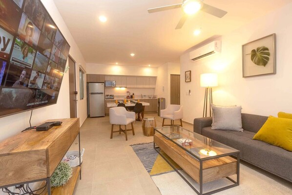 A modern living room with a sofa, coffee table, and TV. There is a kitchen in the background with cabinets and a refrigerator.