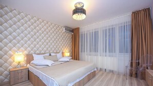 Quarto moderno com cabeceira estofada, cama grande com roupa de cama branca, duas luminárias de cabeceira, mesa de cabeceira de madeira e uma cadeira de plástico transparente.