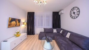 Uma sala de estar moderna com um sofá seccional, uma mesa de centro de vidro com um laptop, um candelabro, um relógio de parede e um quadro na parede.
