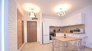 Uma cozinha moderna com armários brancos, porta de madeira, mesa de jantar com cadeiras transparentes e micro-ondas sobre a bancada.