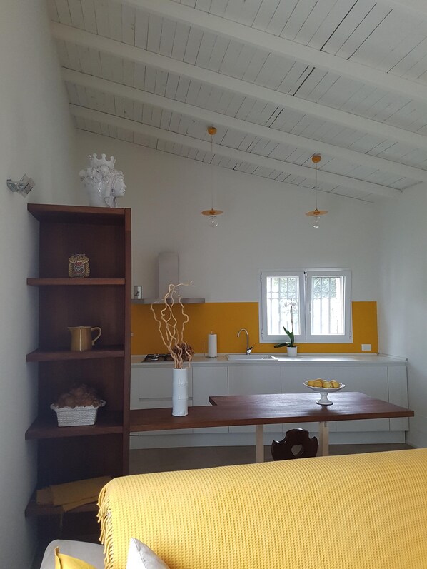 A room with a wooden table, a yellow bedspread, a white vase with branches, a kitchen with a yellow wall, and a window with a view.