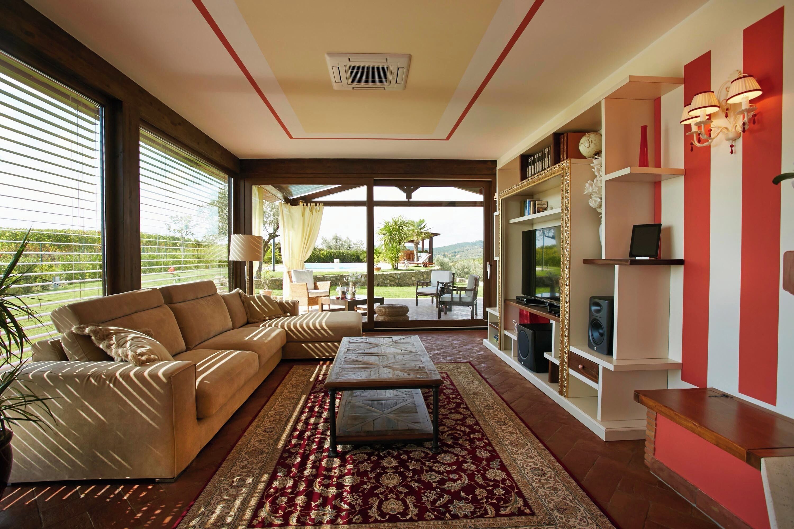 A living room with a beige sofa, a patterned coffee table, and a red and gold rug. There is a bookshelf with decorative items and a red accent wall.