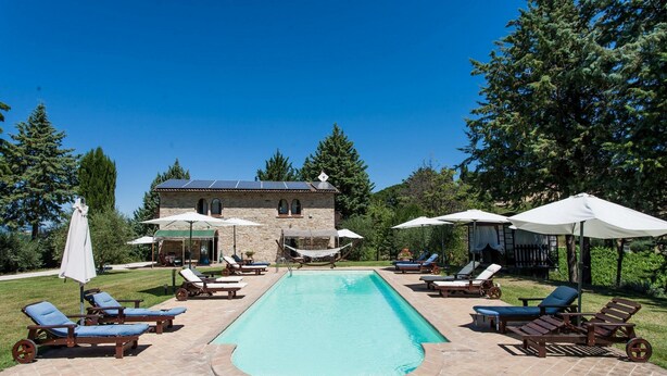 A rectangular swimming pool with lounge chairs and umbrellas, surrounded by a stone patio and a stone building in the background. - Podere Caldaruccio La Pineta - Cottage Umbertide (Perugia)