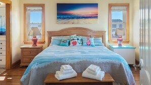 A bedroom with a wooden bed, a painting of a sunset over the ocean, and a view of the beach through the window. - ON THE BEACH! Spiritual Sunrises and Glorious Sunsets, dogs welcome! (Rodanthe)