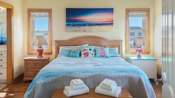 Ein Schlafzimmer mit einem hölzernen Bett, einem Gemälde mit Sonnenuntergang über dem Meer und Blick auf den Strand durch das Fenster.
