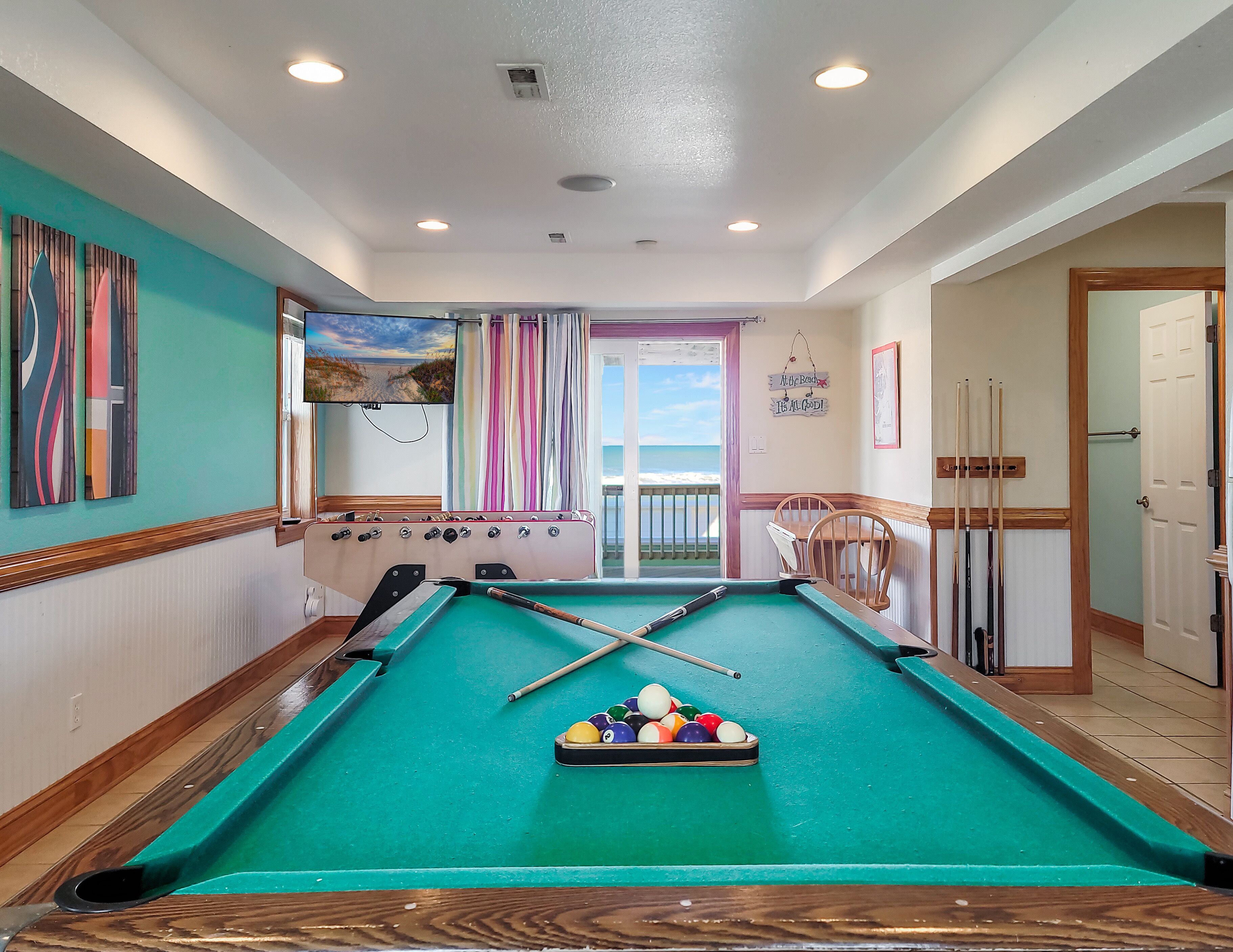A pool table with a green felt surface, a pool cue, and a set of balls. There is a television mounted on the wall, a window with curtains, and a door leading to another room.