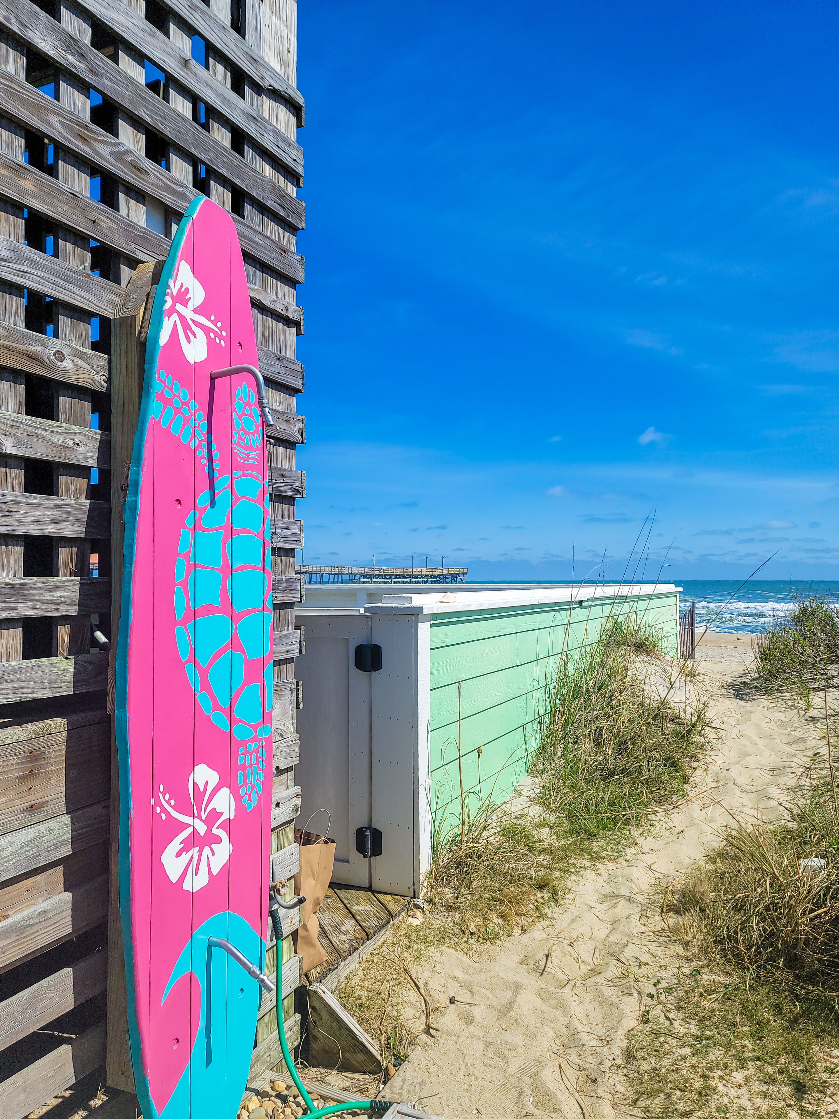 A wooden fence with a colorful surfboard leaning against it, a green building, and a beach with the ocean in the background.