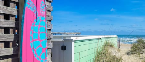 A wooden fence with a colorful surfboard leaning against it, a green building, and a beach with the ocean in the background.