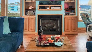 A living room with a fireplace, a television mounted above it, and a view of the beach through the windows. - ON THE BEACH! Spiritual Sunrises and Glorious Sunsets, dogs welcome! (Rodanthe)