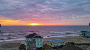 A coastal view with a sunset over the ocean, featuring a multi-story building with balconies and a smaller adjacent structure. - ON THE BEACH! Spiritual Sunrises and Glorious Sunsets, dogs welcome! (Rodanthe)
