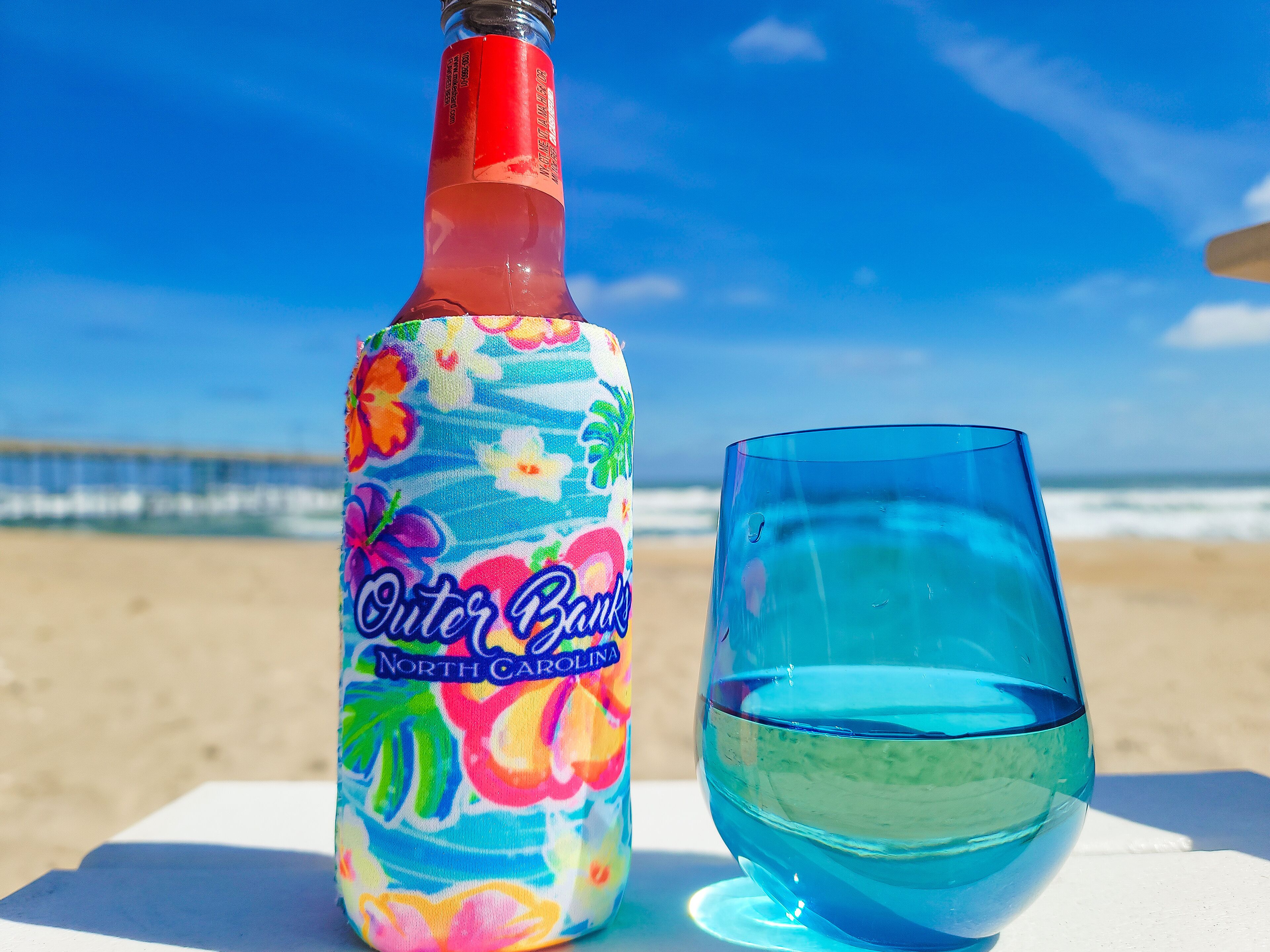 A bottle and glass on a white surface with a beach and ocean in the background.