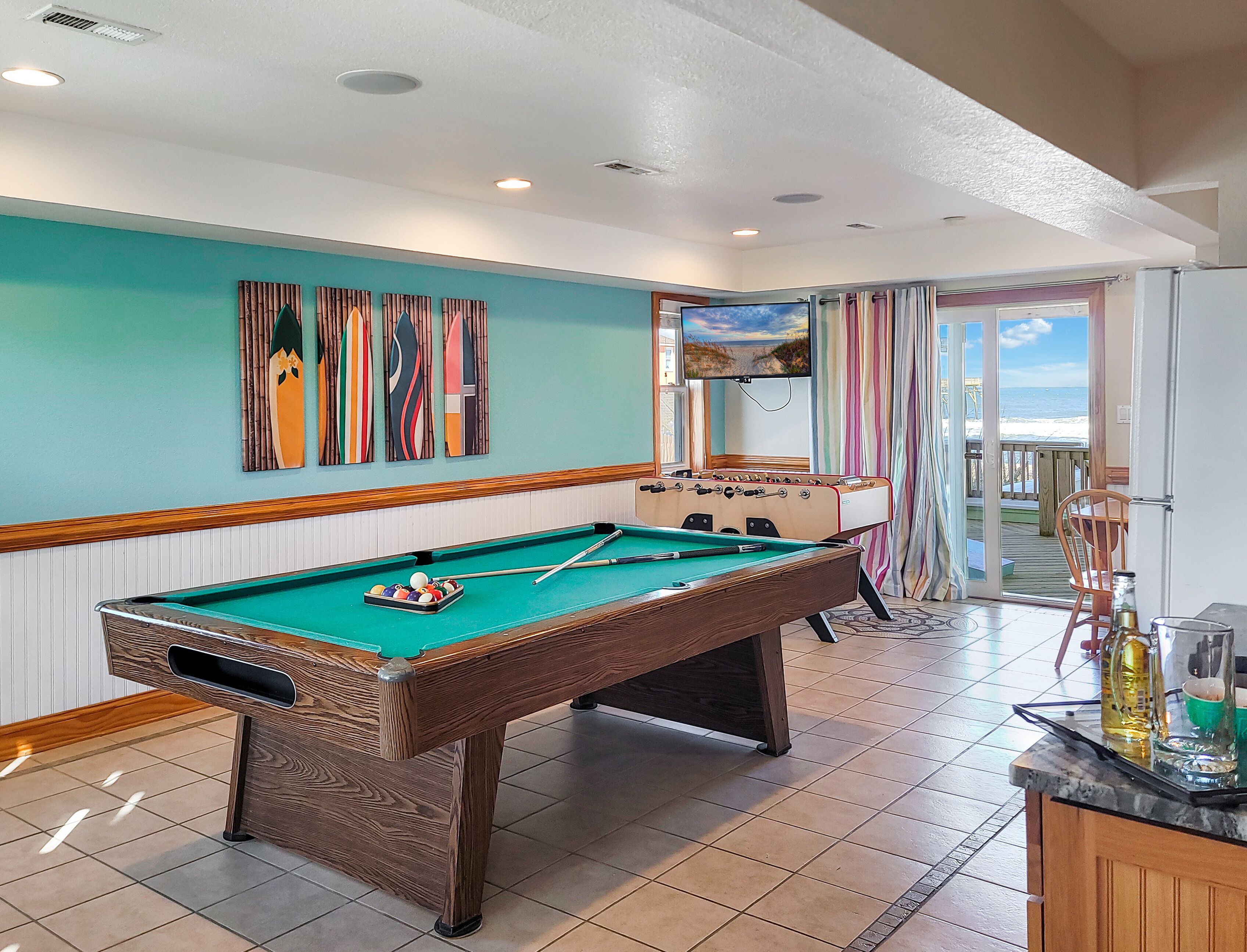 A pool table with a foosball table, a television, and a view of the beach through a glass door.