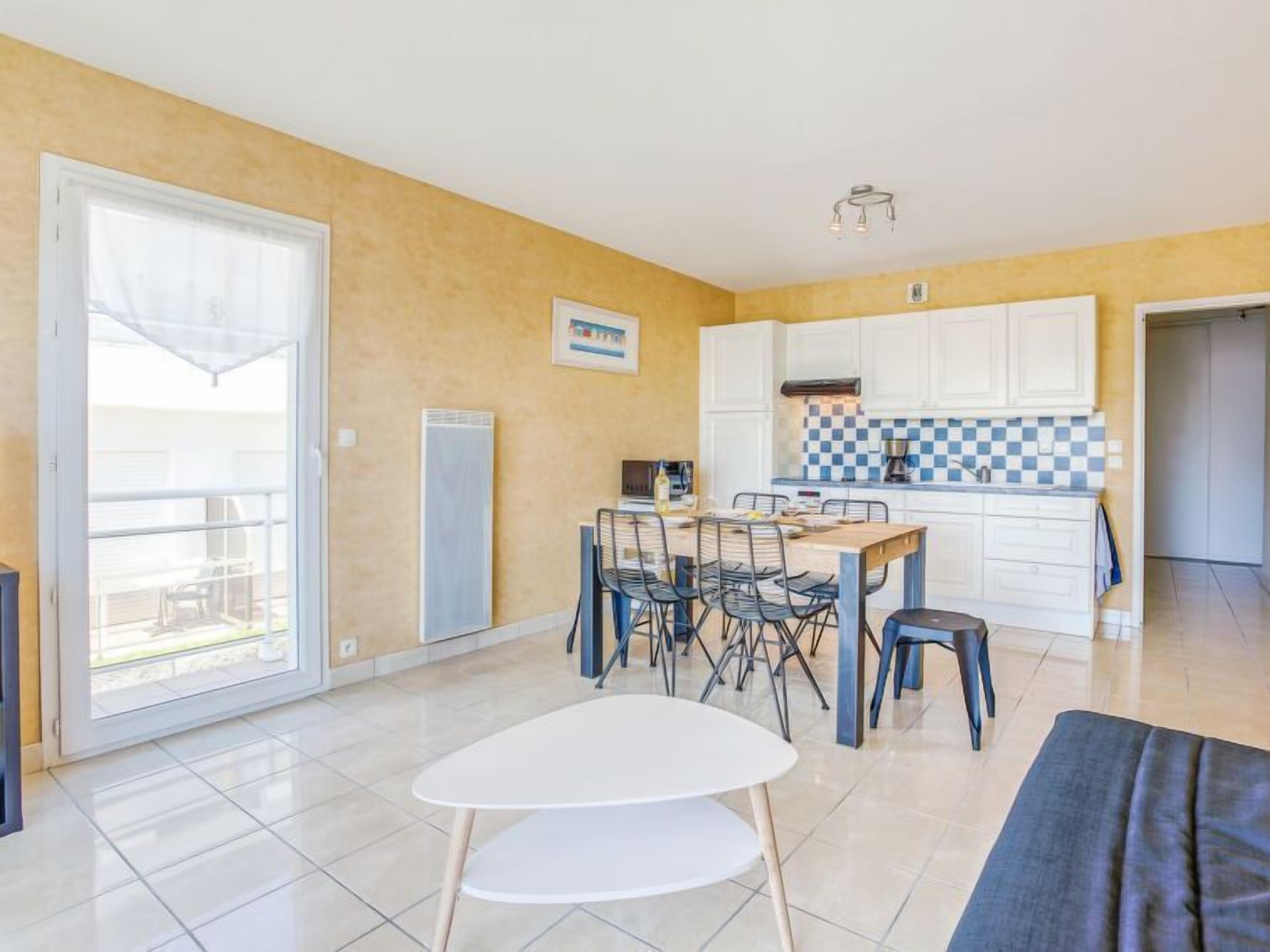 A modern kitchen with white cabinets and a blue and white checkered backsplash. There is a dining area with a white table and chairs, and a large window providing natural light.