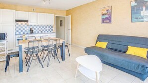 A modern kitchen with white cabinets, a checkered backsplash, and a dining area with a table and chairs. There is a grey sofa with yellow cushions in the living area. - Résidence Ker Eden by Interhome (Quiberon)