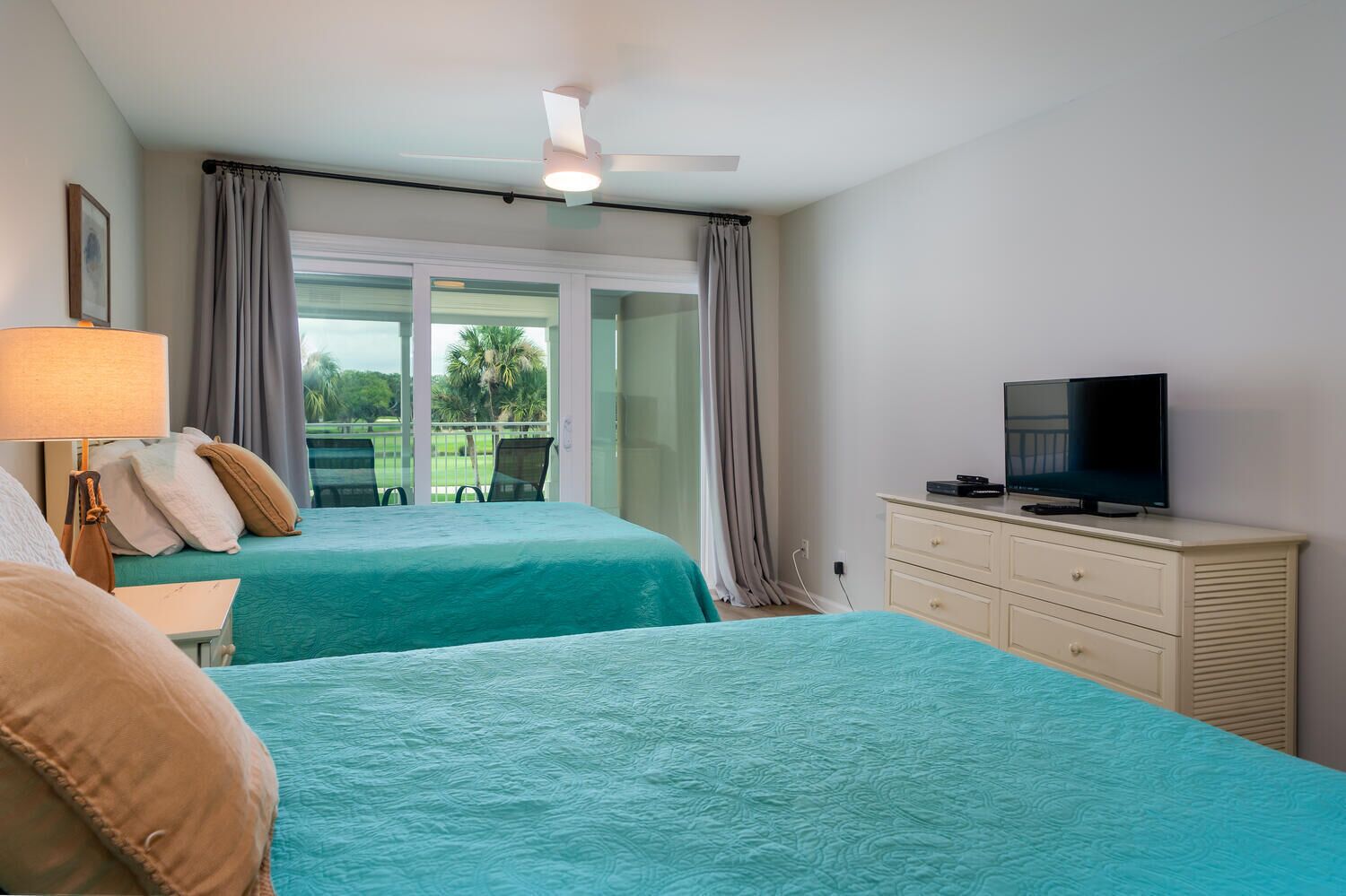 Ein Hotelzimmer mit zwei Betten, einem Fernseher, einem Deckenventilator und Blick auf eine grüne Landschaft.