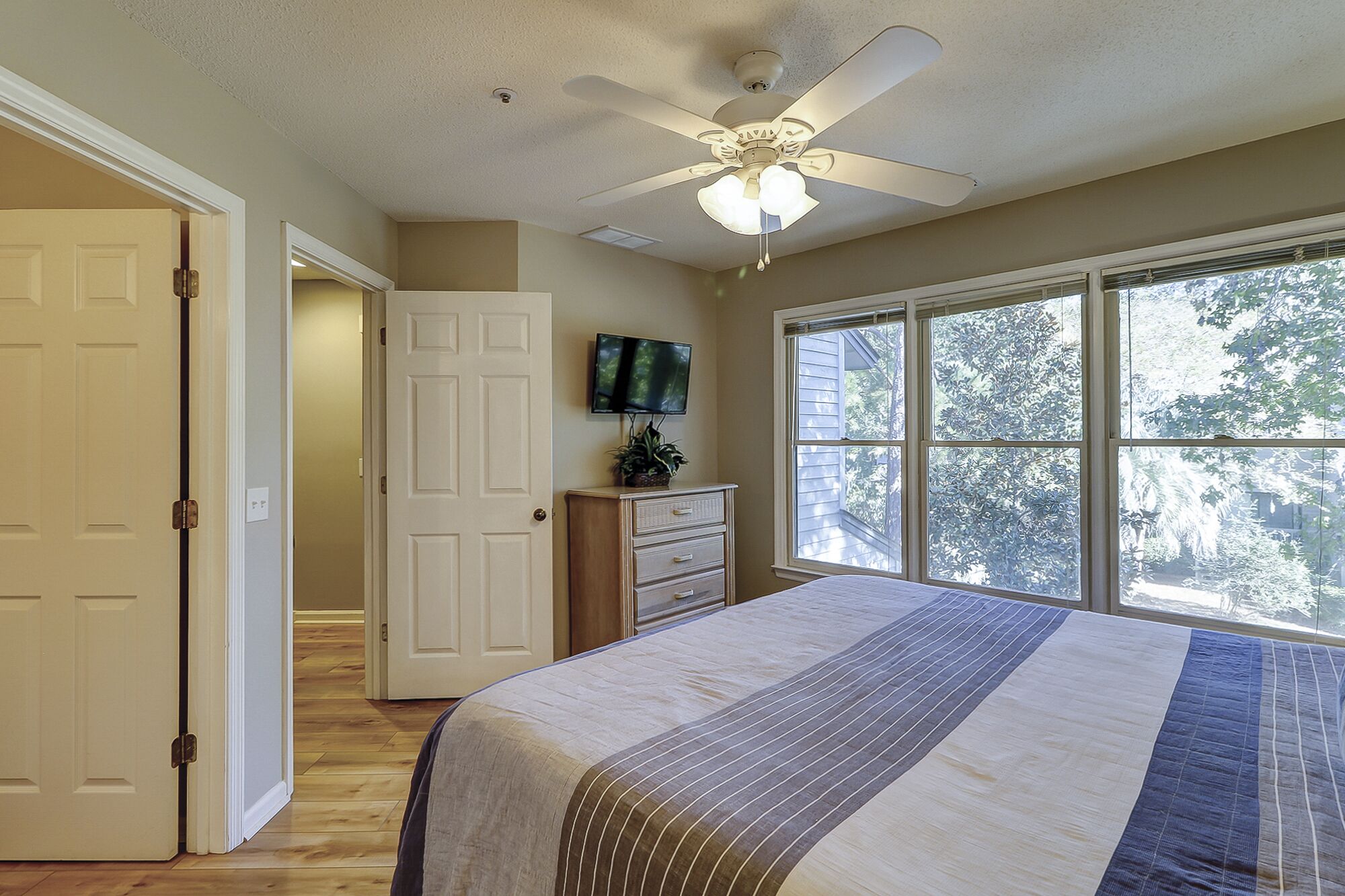 A bedroom with a bed, a ceiling fan, a TV, and a window with a view of trees.