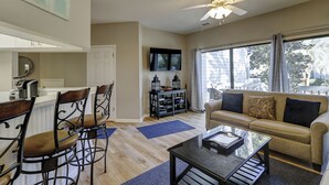 Un salon avec un canapé, une table basse et des fauteuils. Un téléviseur est fixé au mur et un ventilateur de plafond est installé.