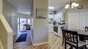 Une cuisine spacieuse avec des meubles blancs, un coin repas comprenant une table et des chaises en bois, et un couloir menant à un salon avec un canapé et un tableau.