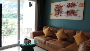 A living room with a brown leather sofa, a glass coffee table, and a painting on the wall. - 6-38 - 2 Bdr Condo With Balcony and Shared Pool (Patong)