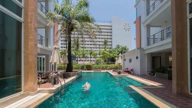 A swimming pool with a person swimming, surrounded by palm trees and a building in the background. - 6-38 - 2 Bdr Condo With Balcony and Shared Pool (Patong)