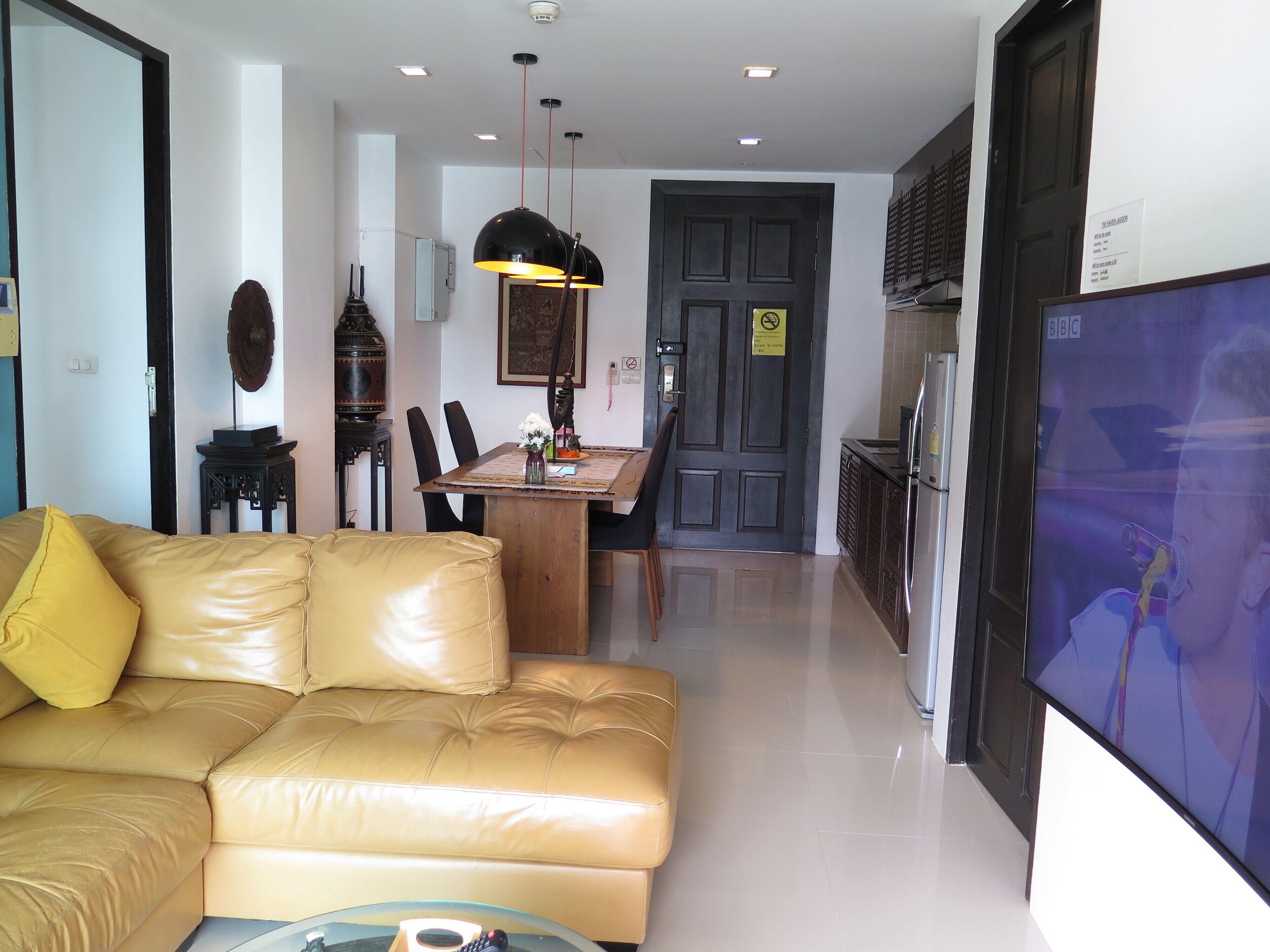 A living room with a leather sofa, a dining area with a table and chairs, a kitchen with a refrigerator, and a TV mounted on the wall.